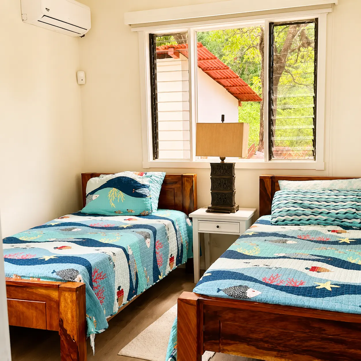 Second bedroom with two twin beds in ocean-themed quilts, AC unit, and window overlooking tropical greenery