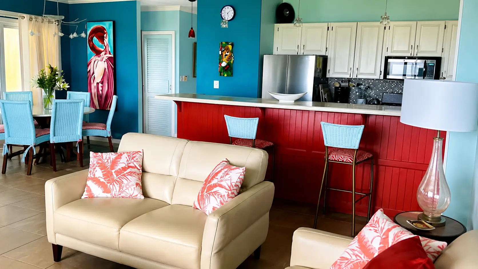 Open-plan living and kitchen with red breakfast bar, blue bar stools, leather seating, and colorful Costa Rican art on the walls