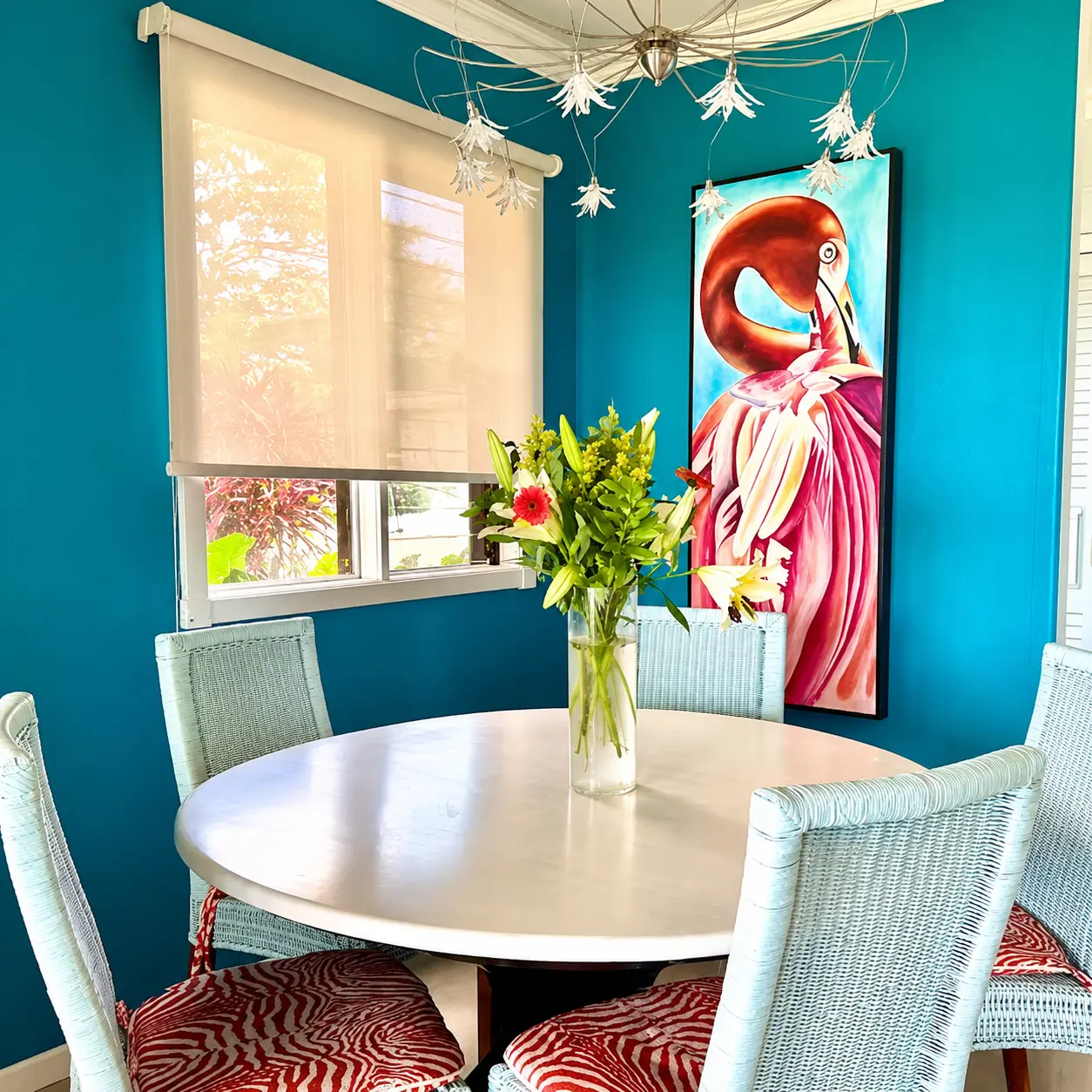 Dining nook with round white table, fresh flowers, wicker chairs with red cushions, flamingo painting, and flower chandelier against teal walls