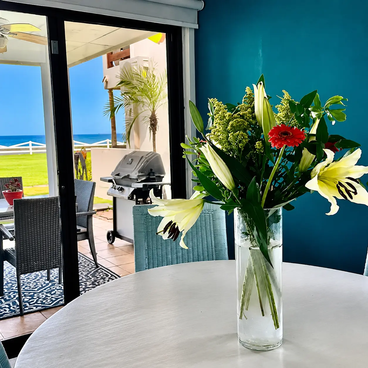 Dining table with fresh lilies looking out through sliding doors to the patio BBQ grill and ocean beyond