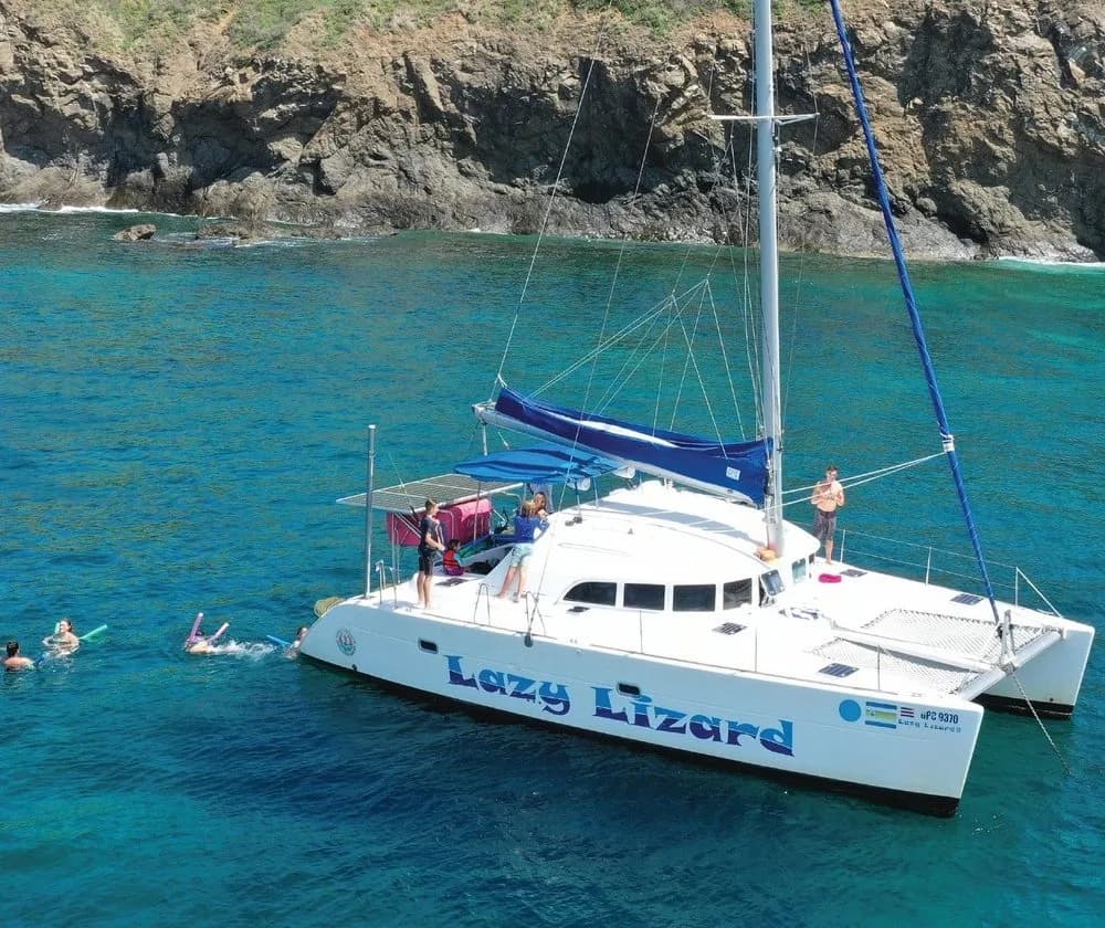 Lazy Lizard catamaran anchored in a turquoise cove with guests snorkeling along rocky cliffs